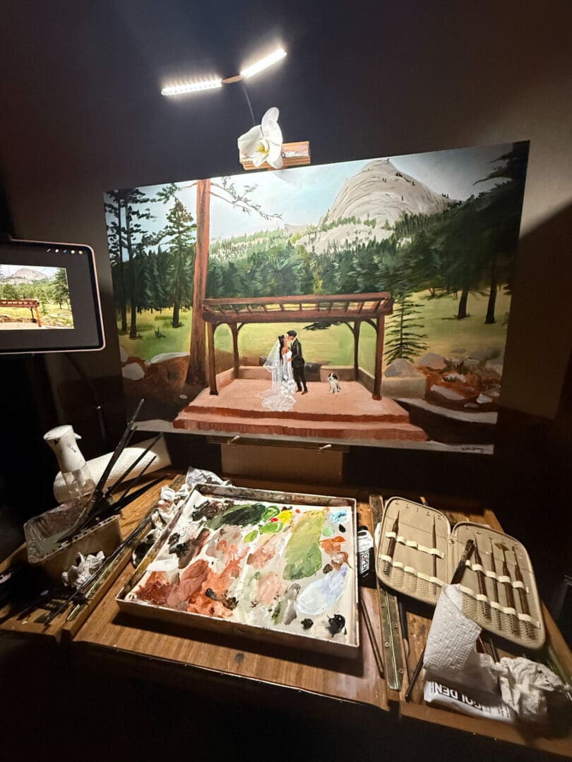 Behind-the-scenes view of a live wedding painting setup on a easel showing a couple kissing under a pergola, with an artist's mixing palette, paintbrushes, and reference tablet in the foreground.