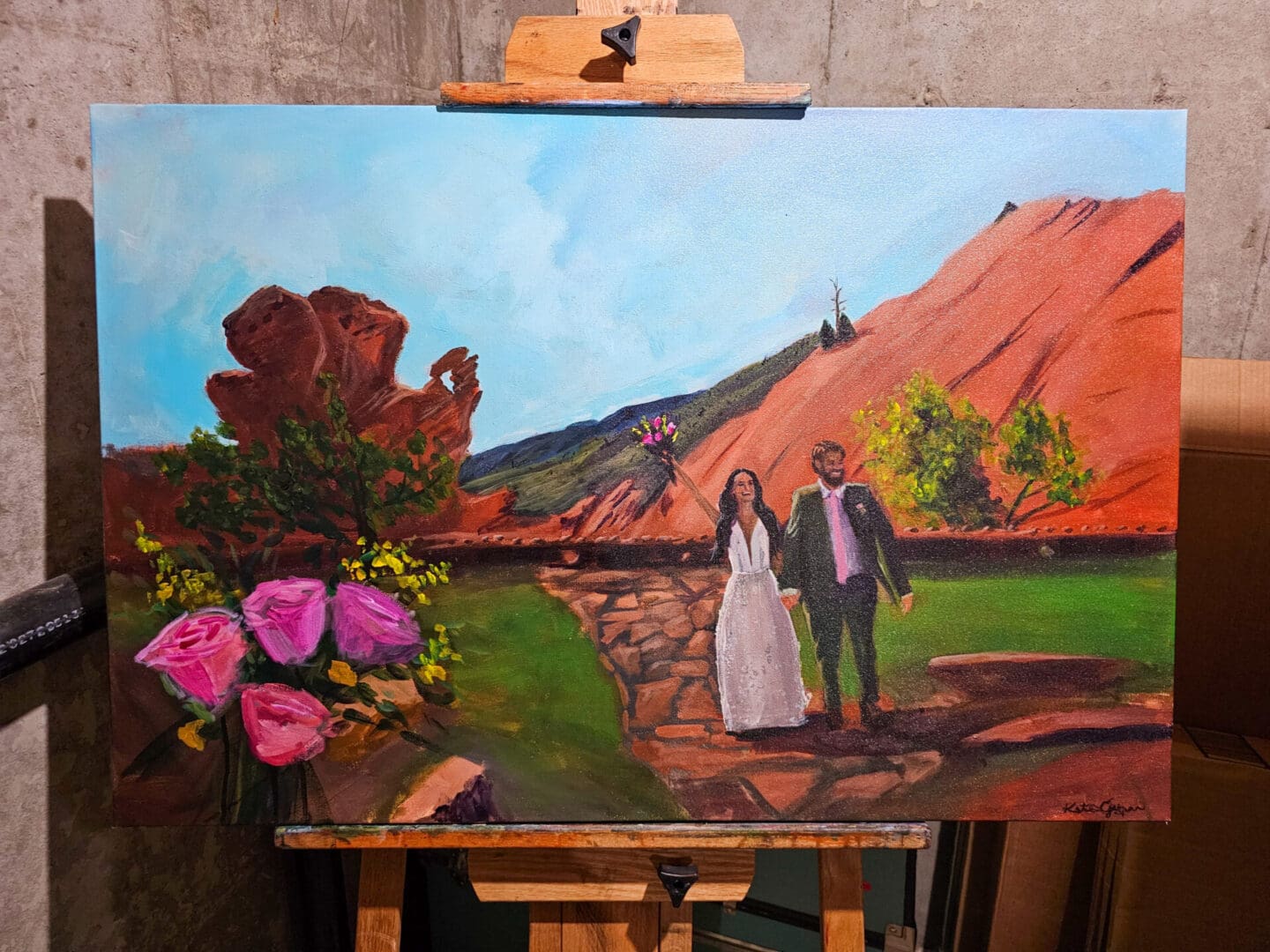 Live wedding painting on an artist easel depicting a bride and groom walking hand-in-hand along a stone path at Red Rocks in Colorado, framed by pink roses and red rock formations.